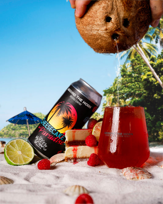A can of 'Cheesecake in Paradise' beer with a colorful tropical design is shown being opened by a person standing on a sandy beach with a coconut in the background, surrounded by beach accoutrements and a glass of the beer's contents is partially visible, indicating the flavor profile of the beer.