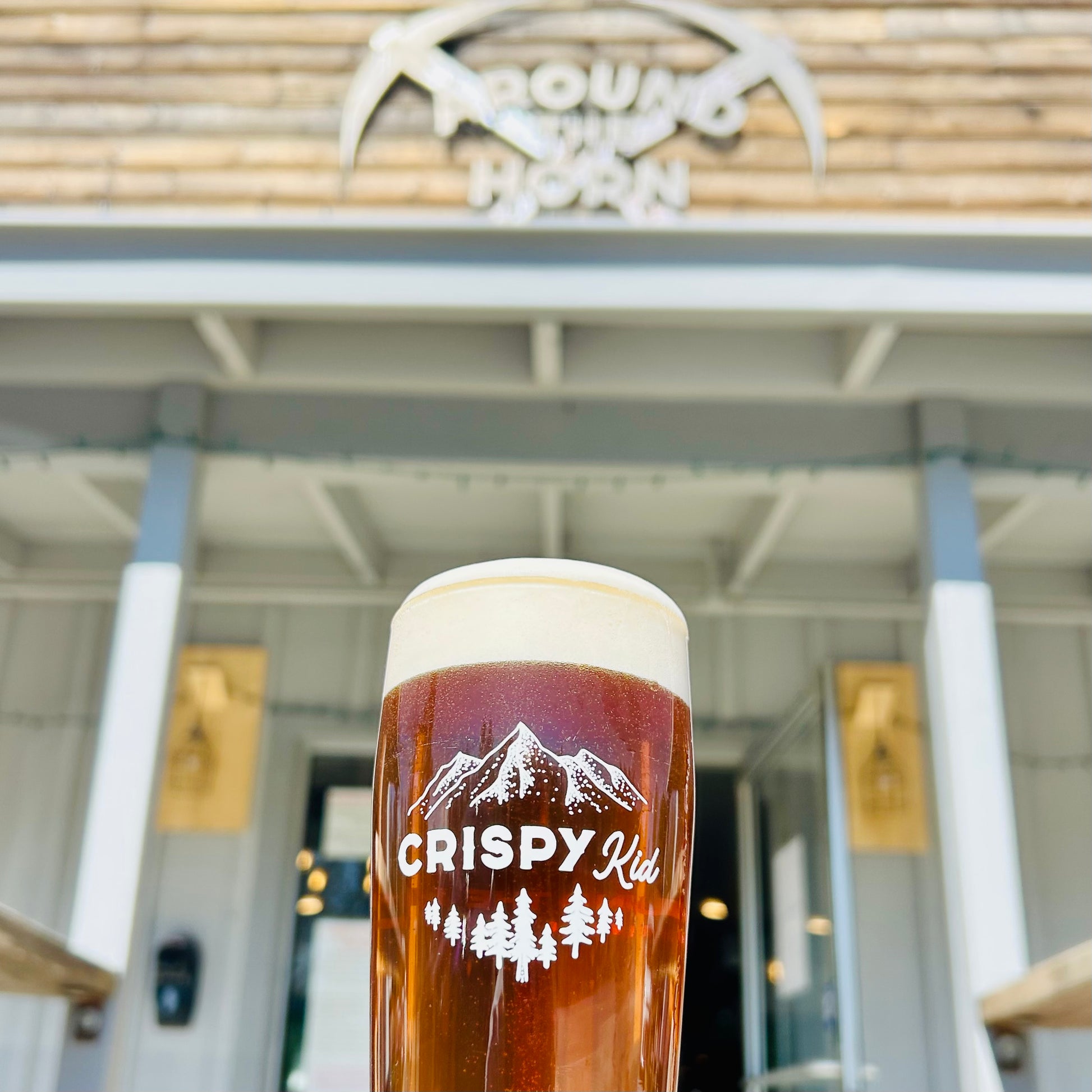Glass of beer with 'Crispy Kid' branding in front of a building with a logo.