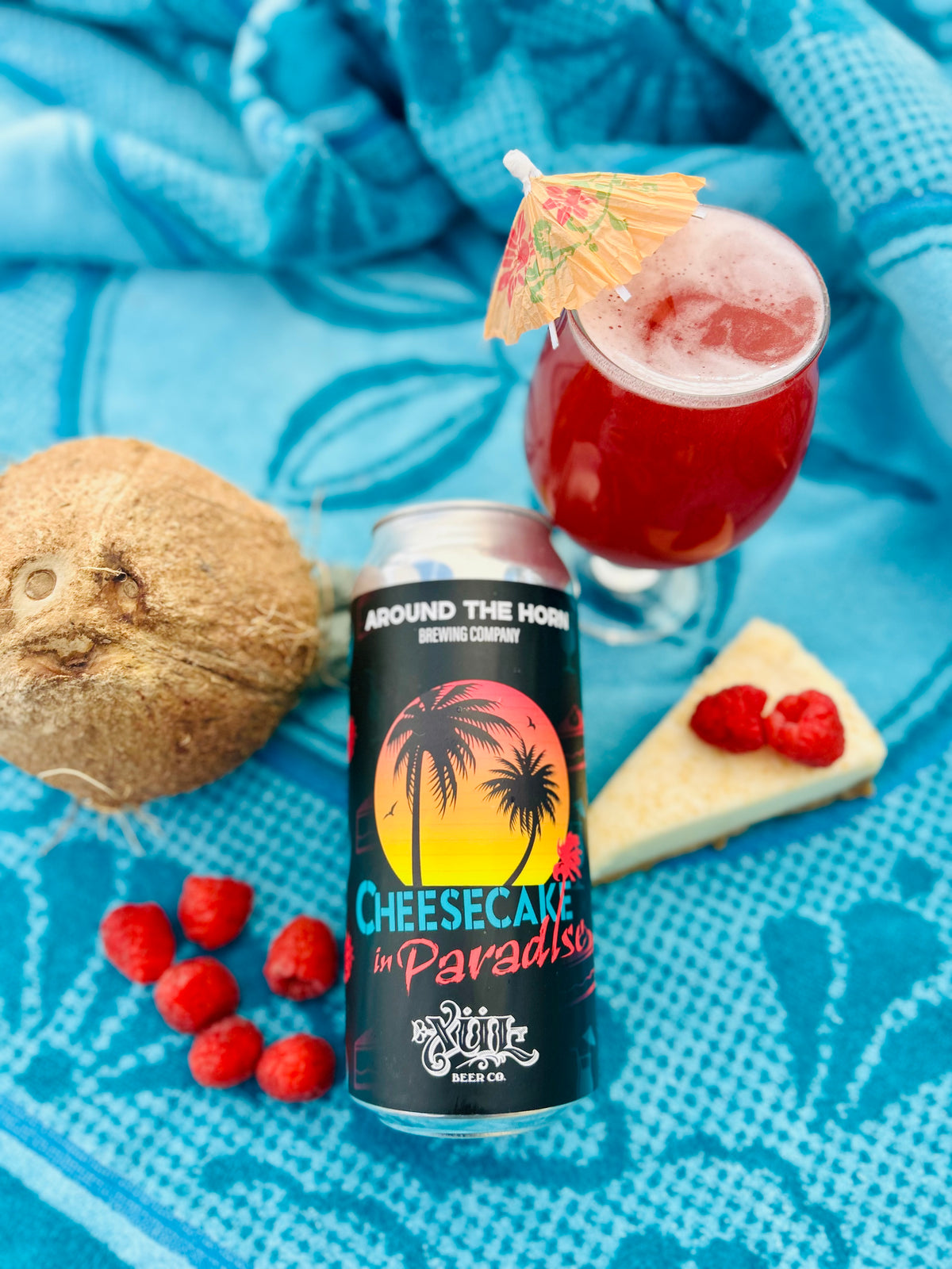 A can of Cheesecake in Paradise beer is styled on a bright blue blanket with a cocktail glass, a coconut, raspberries, and a slice of cake nearby.