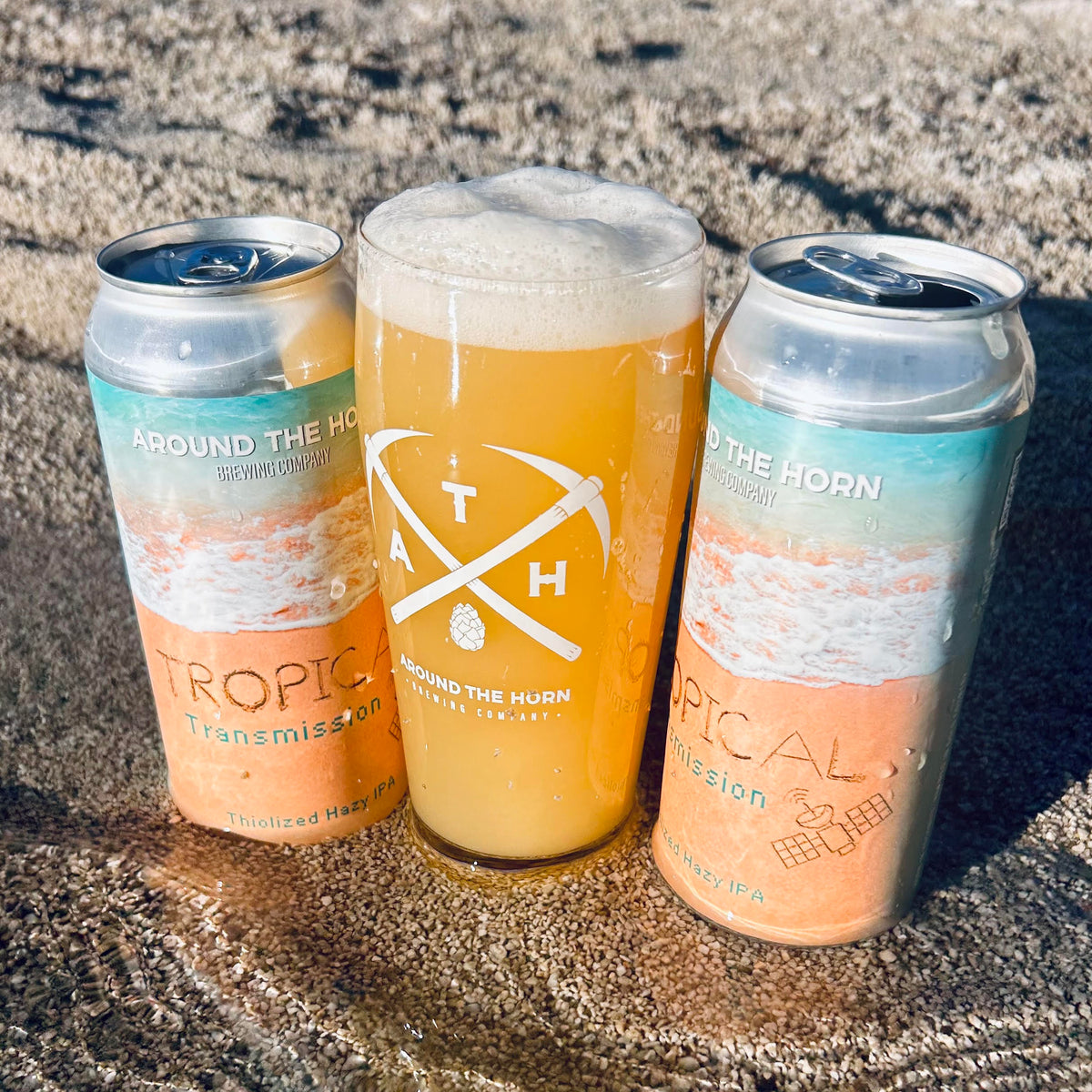 Cans of Around the Horn Brewery Tropical Transmission IPA  next to a glass of golden beer on a sandy beach.