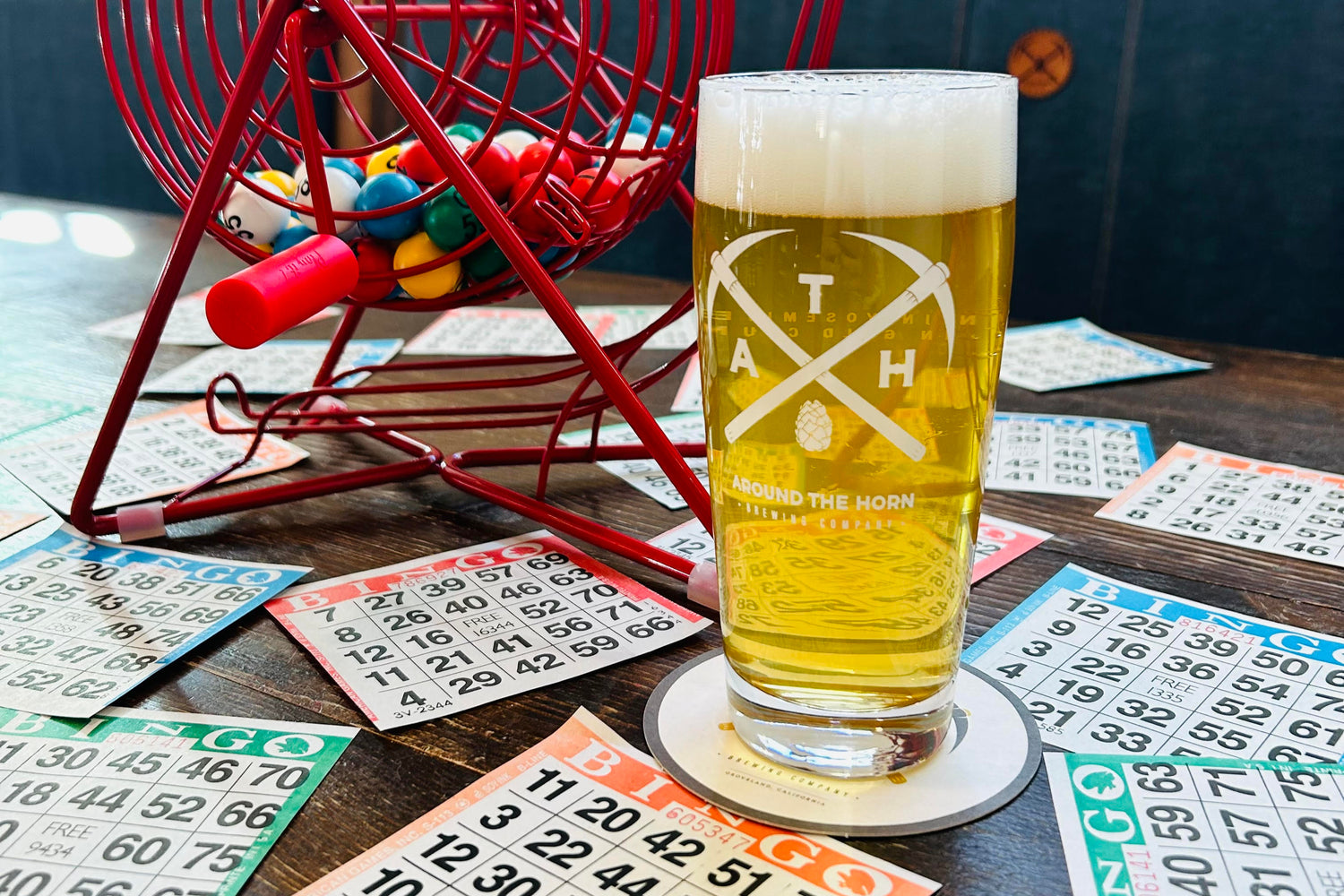Glass of beer with a logo on a table with bingo cards and a red metal wheel.