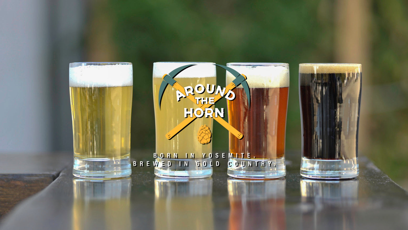 Four flight glasses of different types of beer with a 'Around the Horn' branded logo in the center on a blurred natural background.