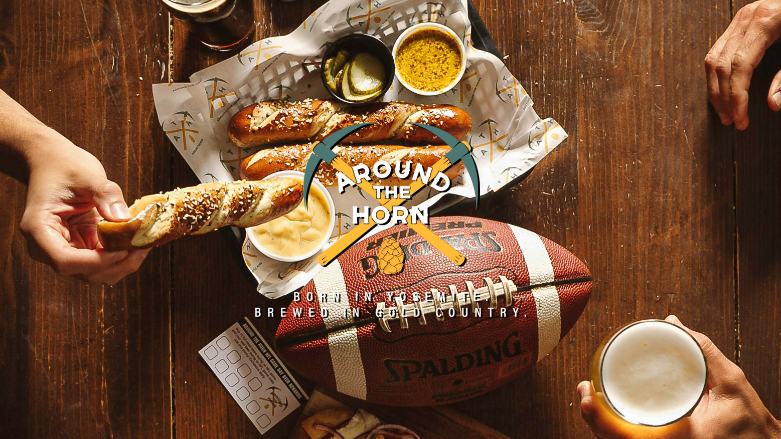 Wooden table with people eating pretzels with mustard and beer cheese and drinking beer. There is a football on the table and there is an Around The Horn logo superimposed on top of the image.