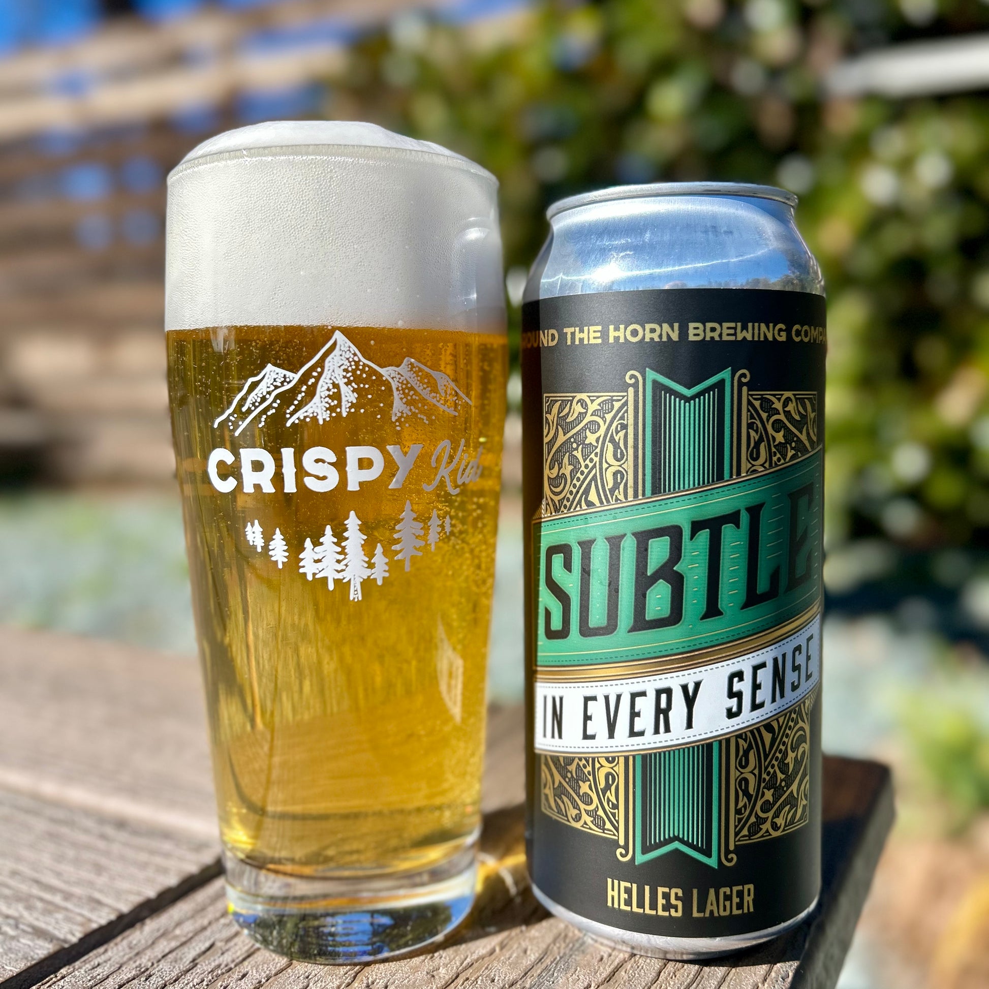 A glass of Helles lager next to a can of Subtle in Every Sense beer on a wood table in a garden.