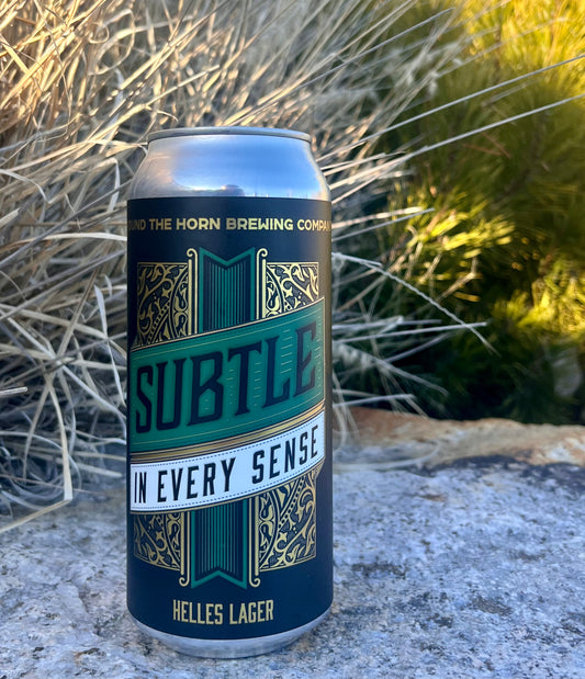 A can of Subtle in Every Sense Helles Lager by Around the Horn Brewing Co., with a green and gold label, placed among dry plants with a background of hills.